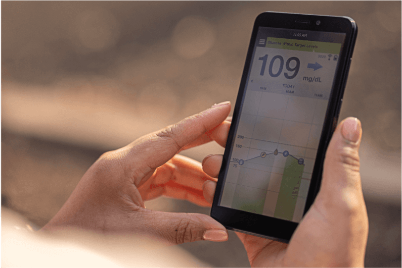 Hands holding a smartphone displaying glucose level chart reading "109 mg/dL" with time stamps in an outdoor setting.