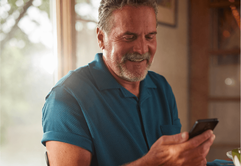 Man smiling, looking at a smartphone, sitting indoors by a window with sunlight streaming through.