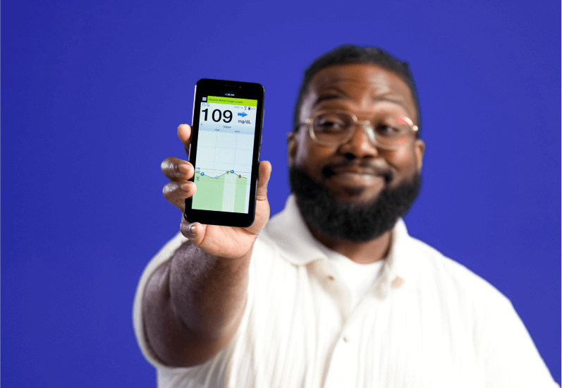 Man holds smartphone displaying "109 mg/dL" glucose reading graph, smiling, standing against solid blue background.